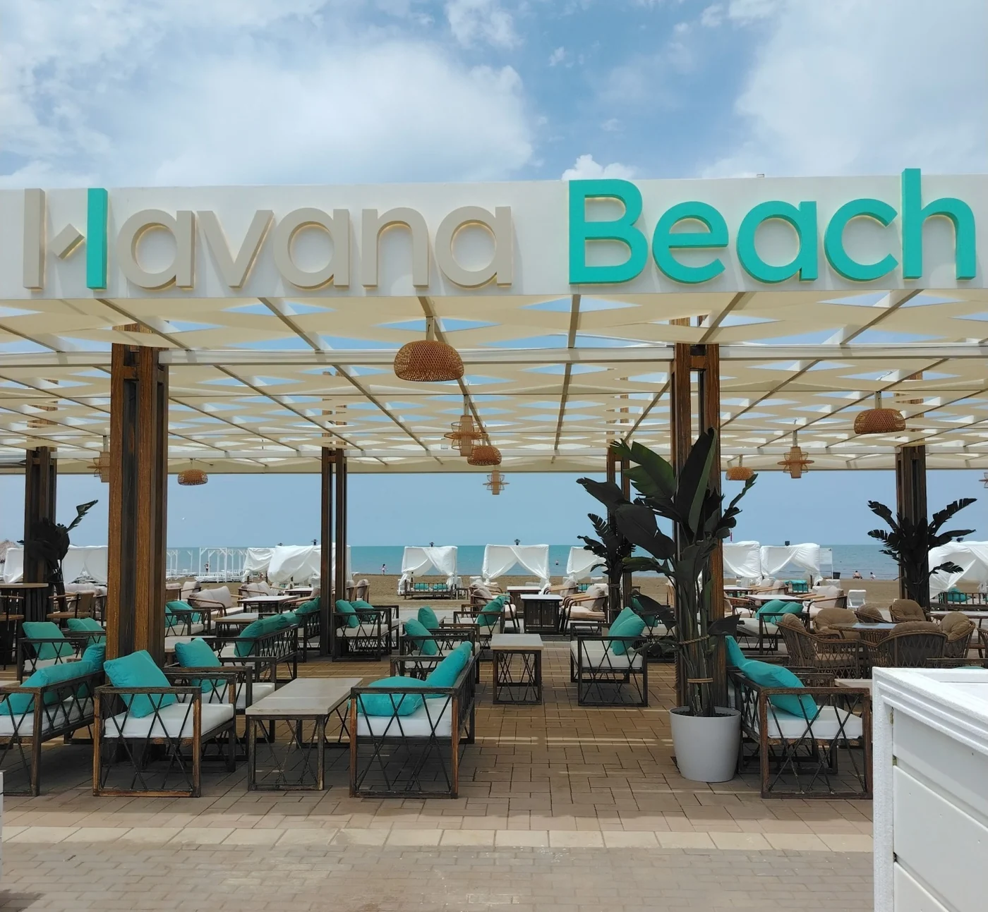 Вход в Havana Beach: белая балка с объёмными буквами Havana Beach над деревянной перголой, бирюзовые подушки на лаунж-диванах, за ними — белые кабаны и Каспий